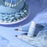 Colour Mill Baby Blue Oil Blend bottle beside blue icing piping bags and a decorated cake