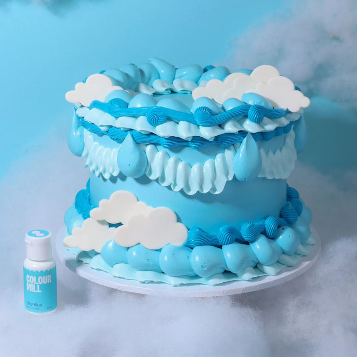 Blue piped cake with white cloud decorations beside Colour Mill Sky Blue food colour